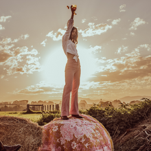 Load image into Gallery viewer, Wandering Folk Woven Throw Rosé | Boho ; Farbe: Rosa, Ockergelb ; mit Fransen ; Blumenmuster ; Lifestyle Foto auf Heuballen stehendes Model mit Glas in der Hand ; Campingdecke